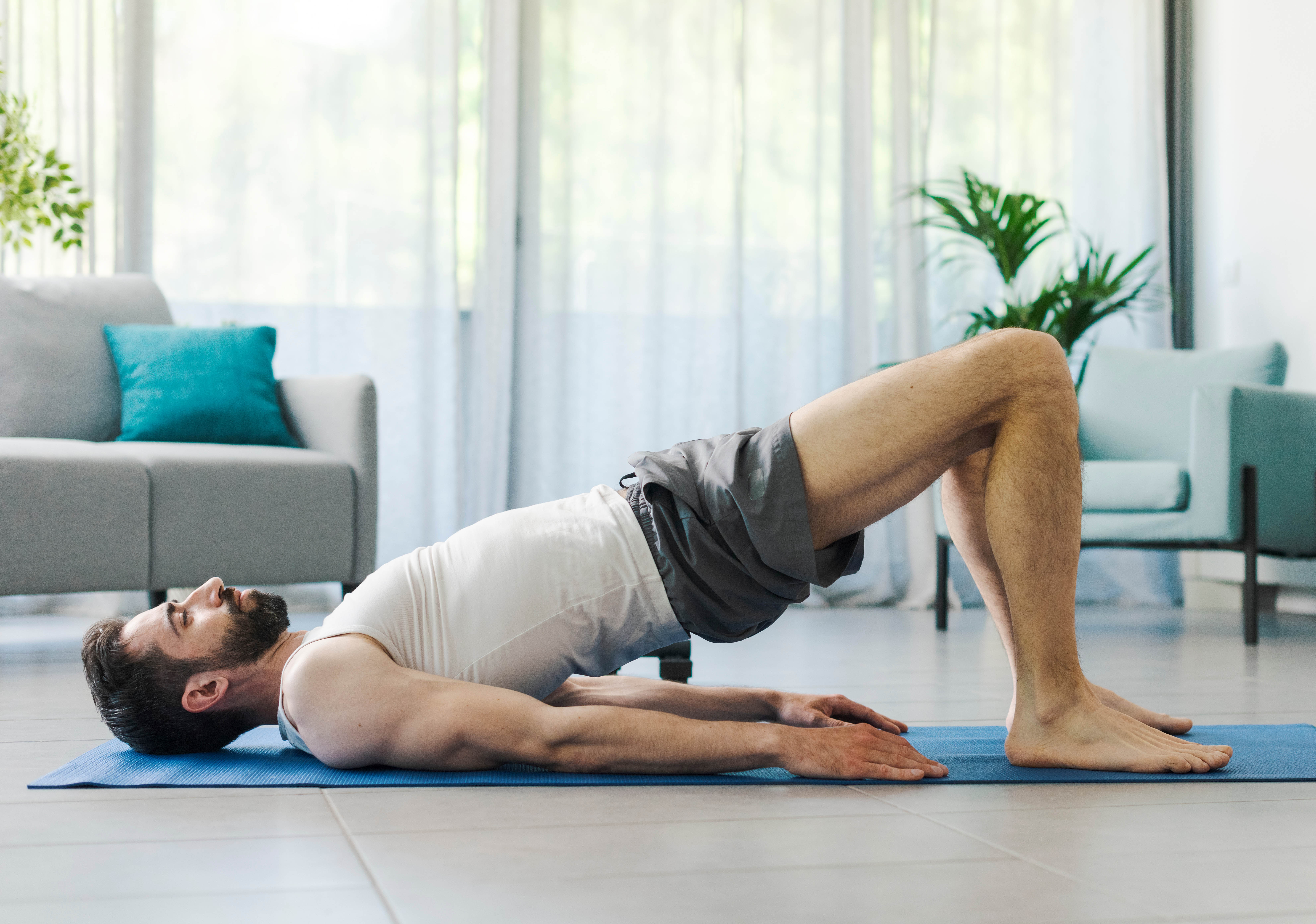 Athletic man in a bridge pose on a yoga mat in his living room; couch, chair, and plants in the background Athletic man in a bridge pose on a yoga mat in his living room; couch, chair, and plants in the background